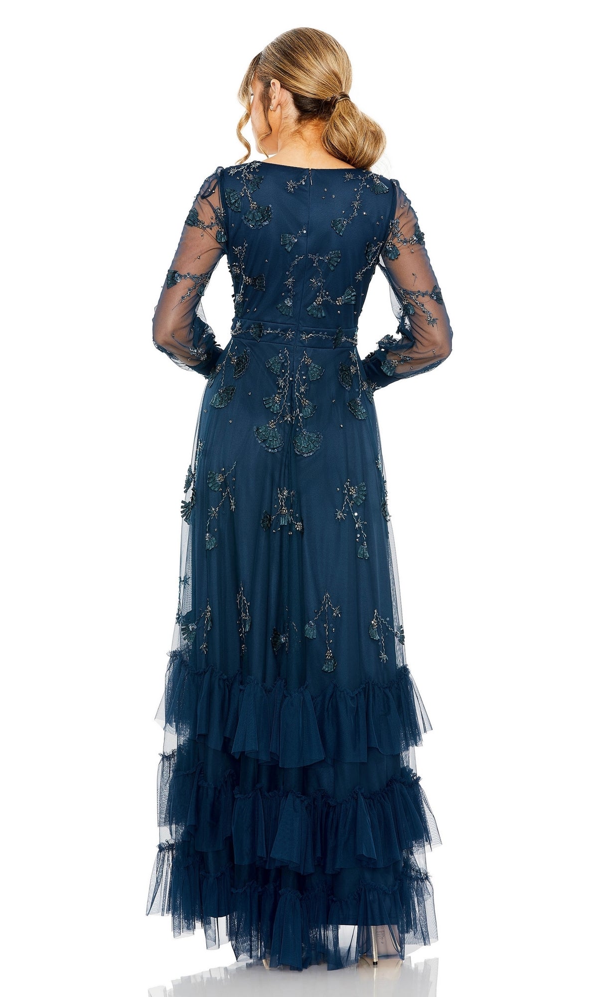 A woman stands with her back to the camera, wearing the Mac Duggal 9256—a long sleeve blue formal dress featuring sheer fabric, floral embroidery, and tiered ruffle layers.